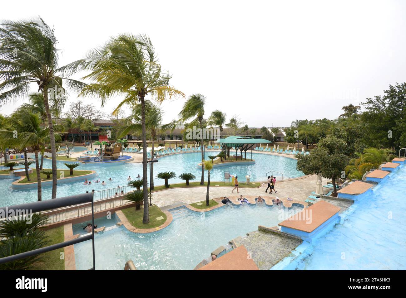 Tourists at a luxury resort pool with waterfall and sun loungers in the background on a cloudy blue sky day. Wide-angle panoramic view. Winter in Bra Stock Photo