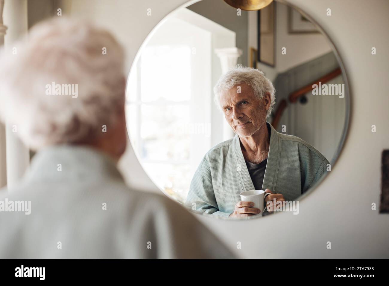 Senior man holding coffee cup while looking in mirror at home Stock Photo