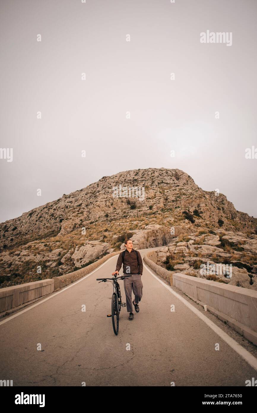 Young man wheeling bicycle on road against mountain Stock Photo