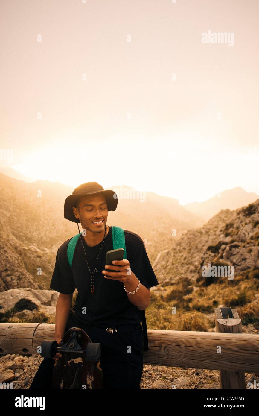 Smiling young man using mobile phone while sitting on railing against sky Stock Photo