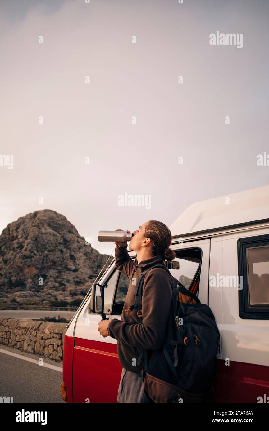 Young man drinking water while standing near van on vacation Stock Photo