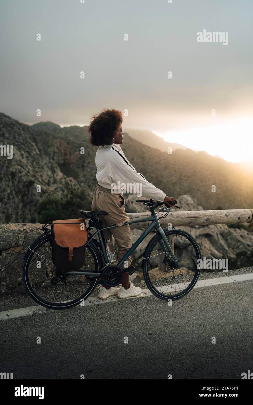 Woman exploring mountains while standing with cycle on road at sunset Stock Photo