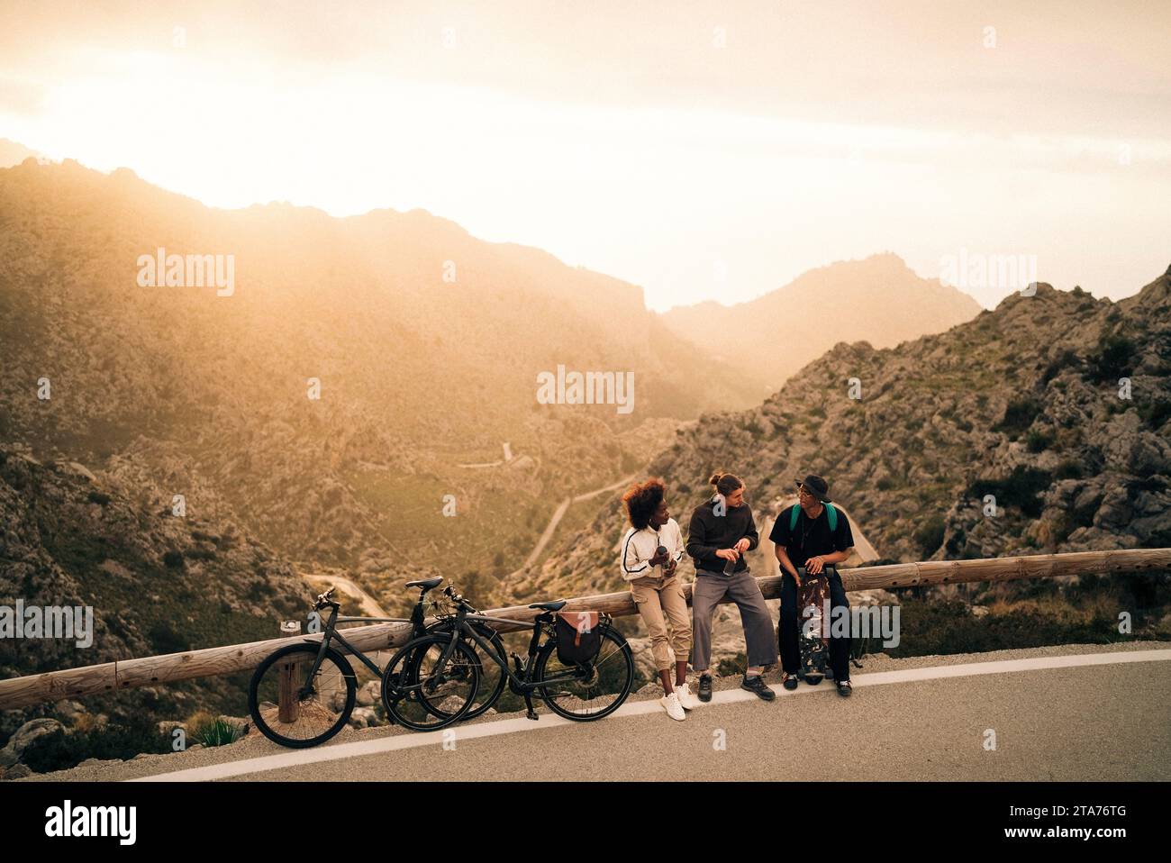 Male and female explorers with bicycles talking while sitting on railing at sunset Stock Photo