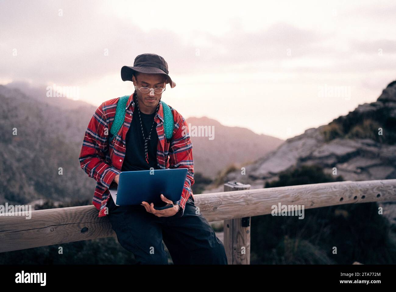 Young man using laptop while sitting on railing at sunset Stock Photo