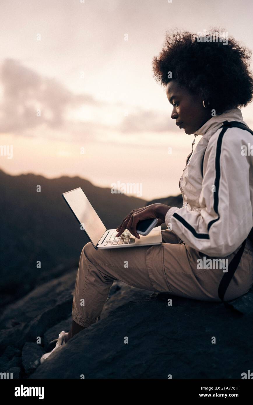 Woman with curly hair using laptop while sitting on rock at sunset Stock Photo