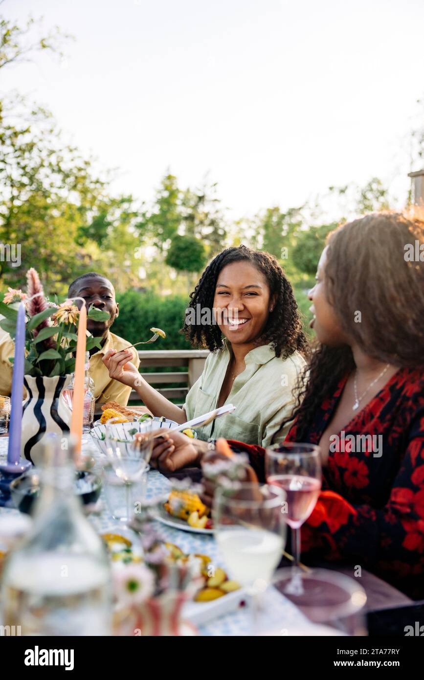 Smiling woman having food with friends sitting at dining table during party in back yard Stock Photo
