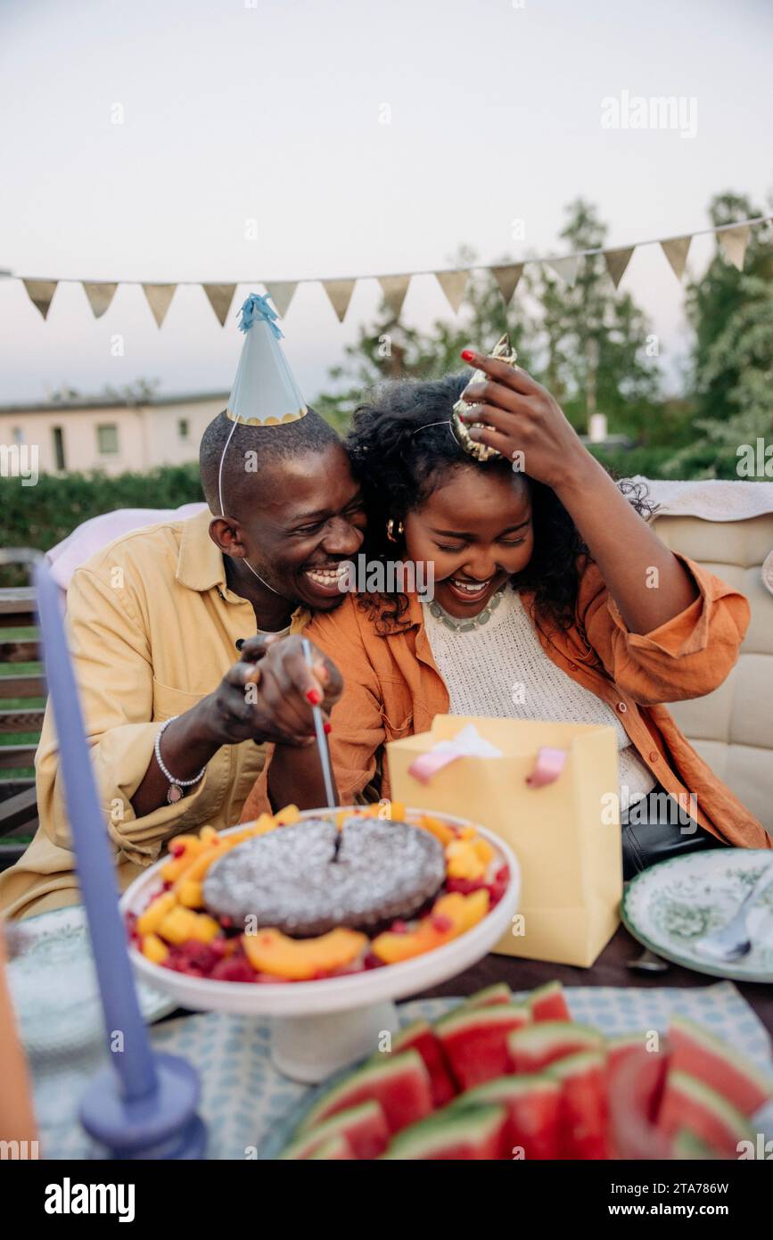 Happy young man sitting with female friend cutting cake while celebrating birthday in back yard Stock Photo