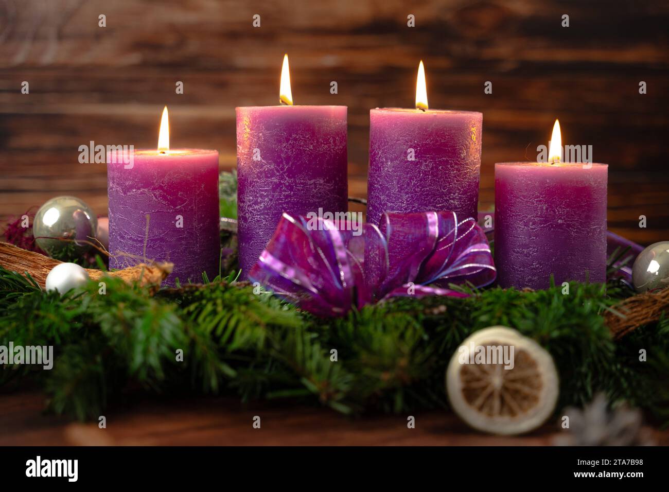 November 24, 2023: Advent wreath with four burning purple candles for the fourth Sunday of Advent in front of a rustic wooden background *** Adventskranz mit vier brennenden Lila Kerzen zum vierten