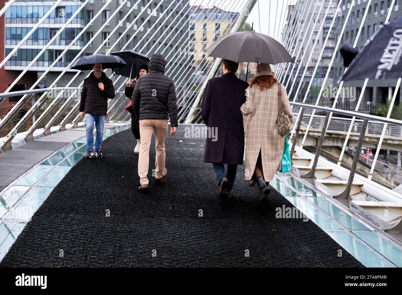 Five people walk on a rainy day across the Zubizurri Bridge, one of the most famous bridges in Bilbao, Spain. Stock Photo