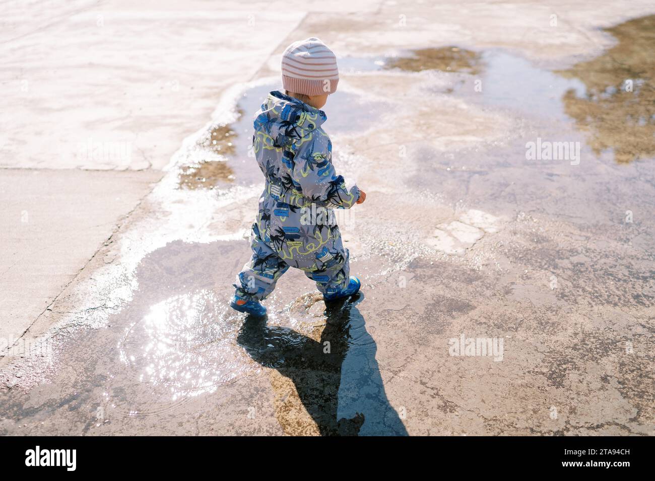 Little girl in rubber boots walks through a puddle on the pavement ...