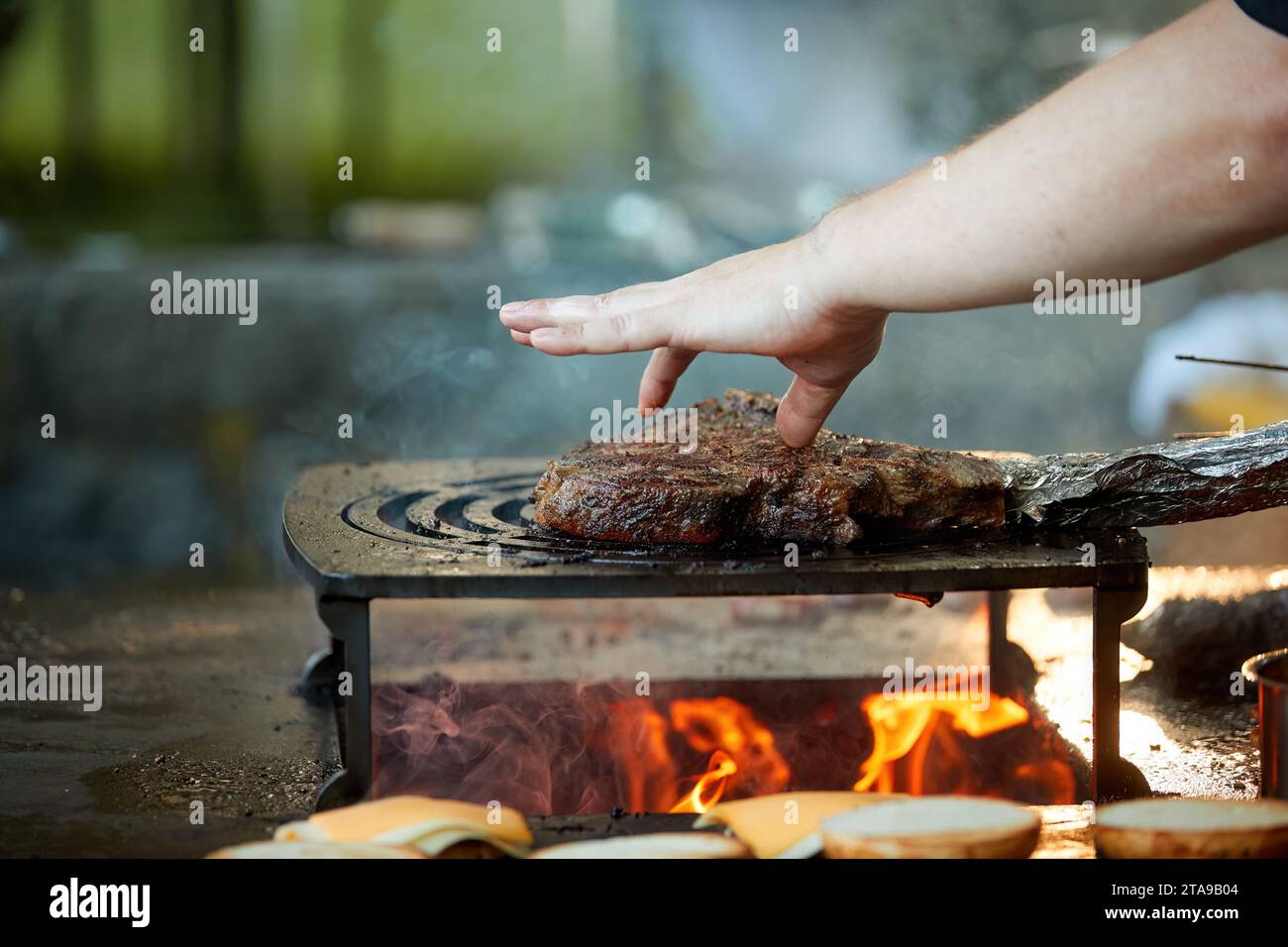 A chef controls a steak on an outdoor grill by finger. Close up of cooking at garden festival, flames, shallow depth of field, very colorful blurred b Stock Photo