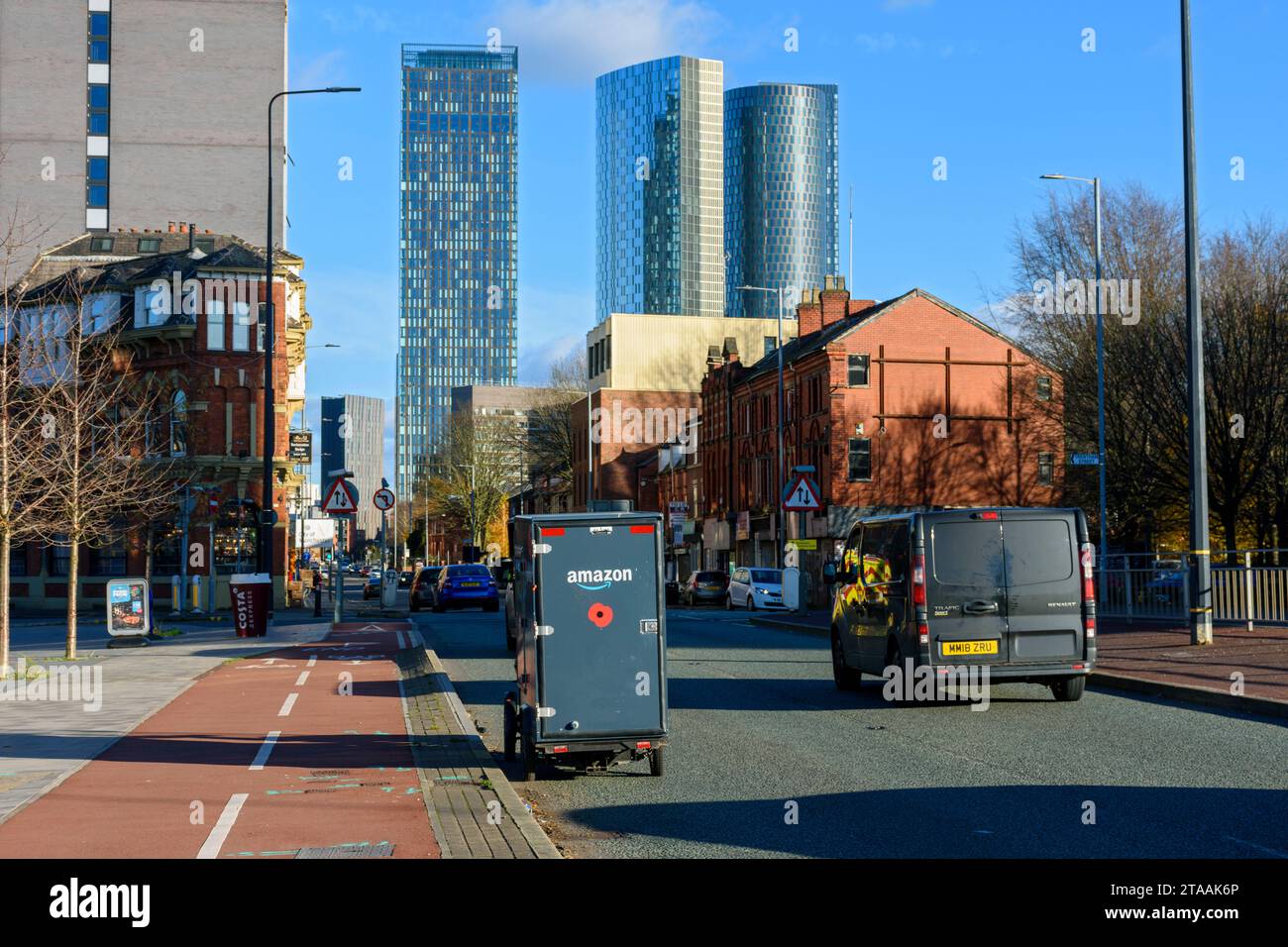An Amazon e-cargo delivery vehicle on Chester Road, Manchester, England, UK. The Elizabeth Tower, The Blade and the Three60 apartment blocks behind. Stock Photo