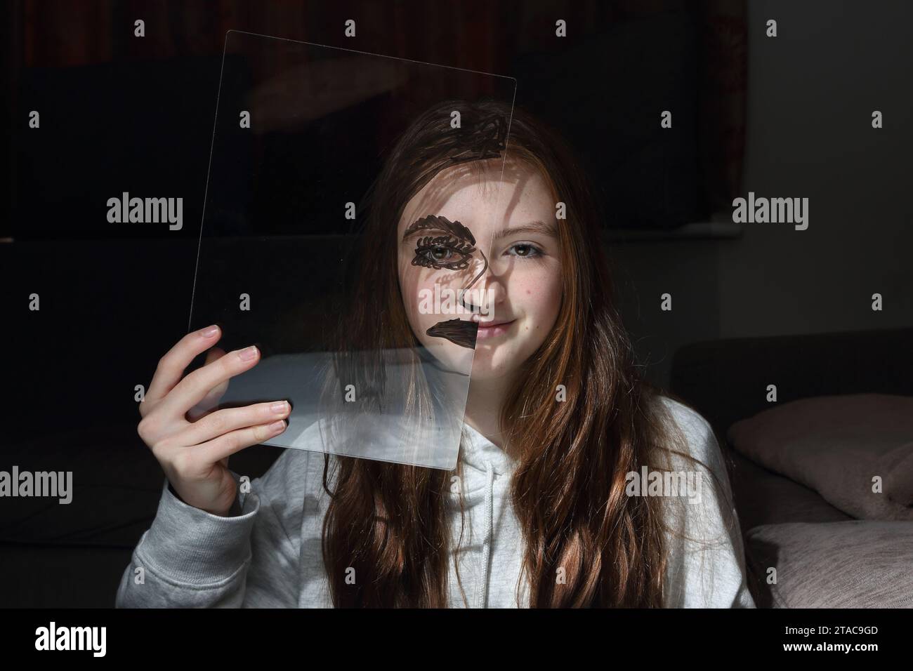 Teenage girl holds transparent plastic board in front of her face with half a face drawn on it - eye and half of mouth Stock Photo