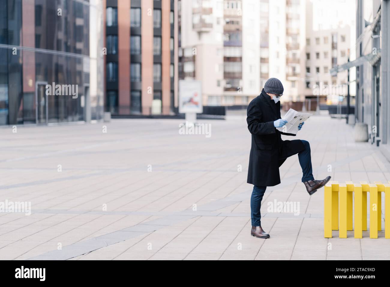Man in a coat reading a newspaper on a city street, wearing a mask and blue gloves Stock Photo