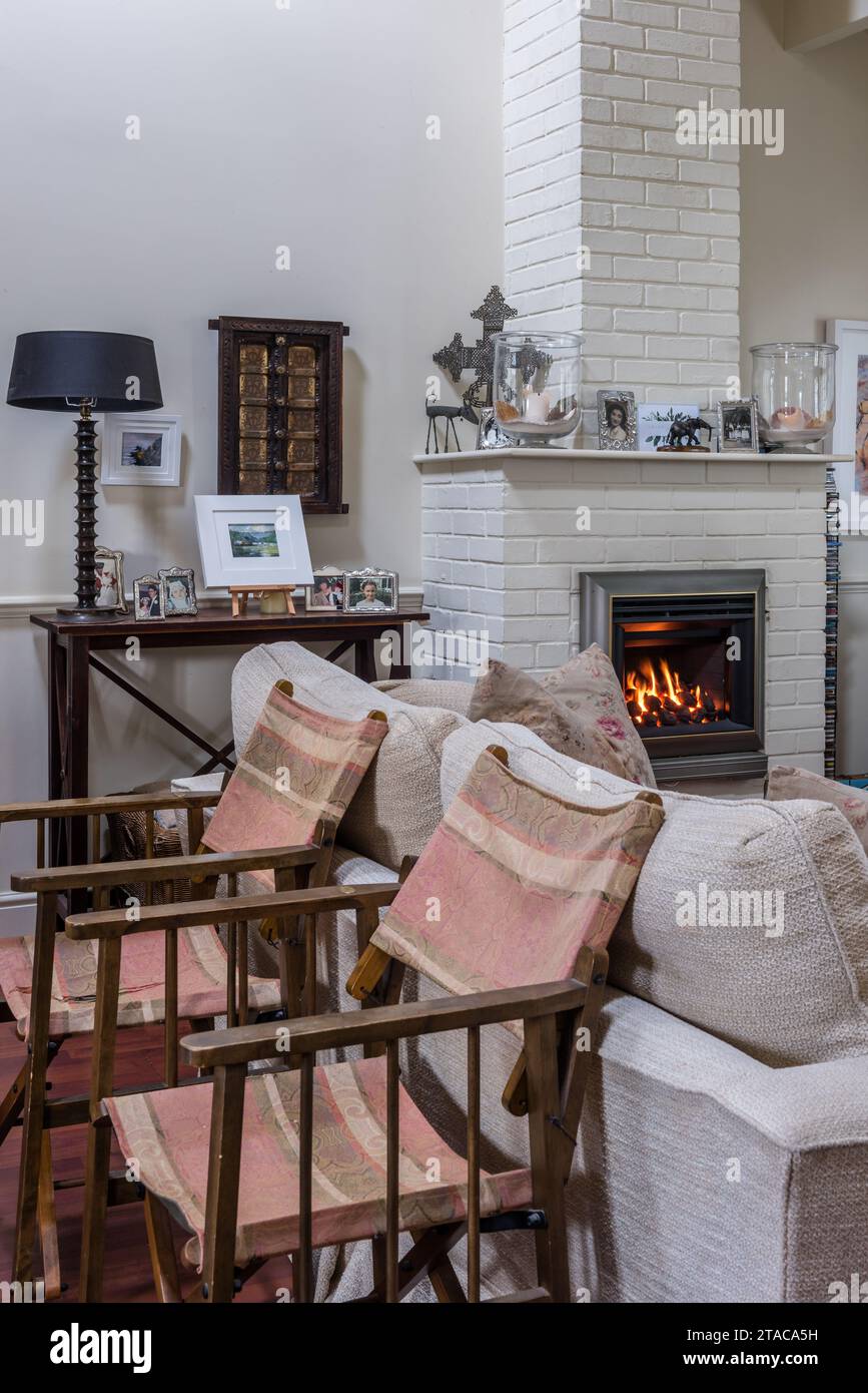 Painted brick fireplace with 'real flame' gas and director's chairs by Andrew Martin in colonial style single-storey Truro home, Cornwall, UK. Stock Photo