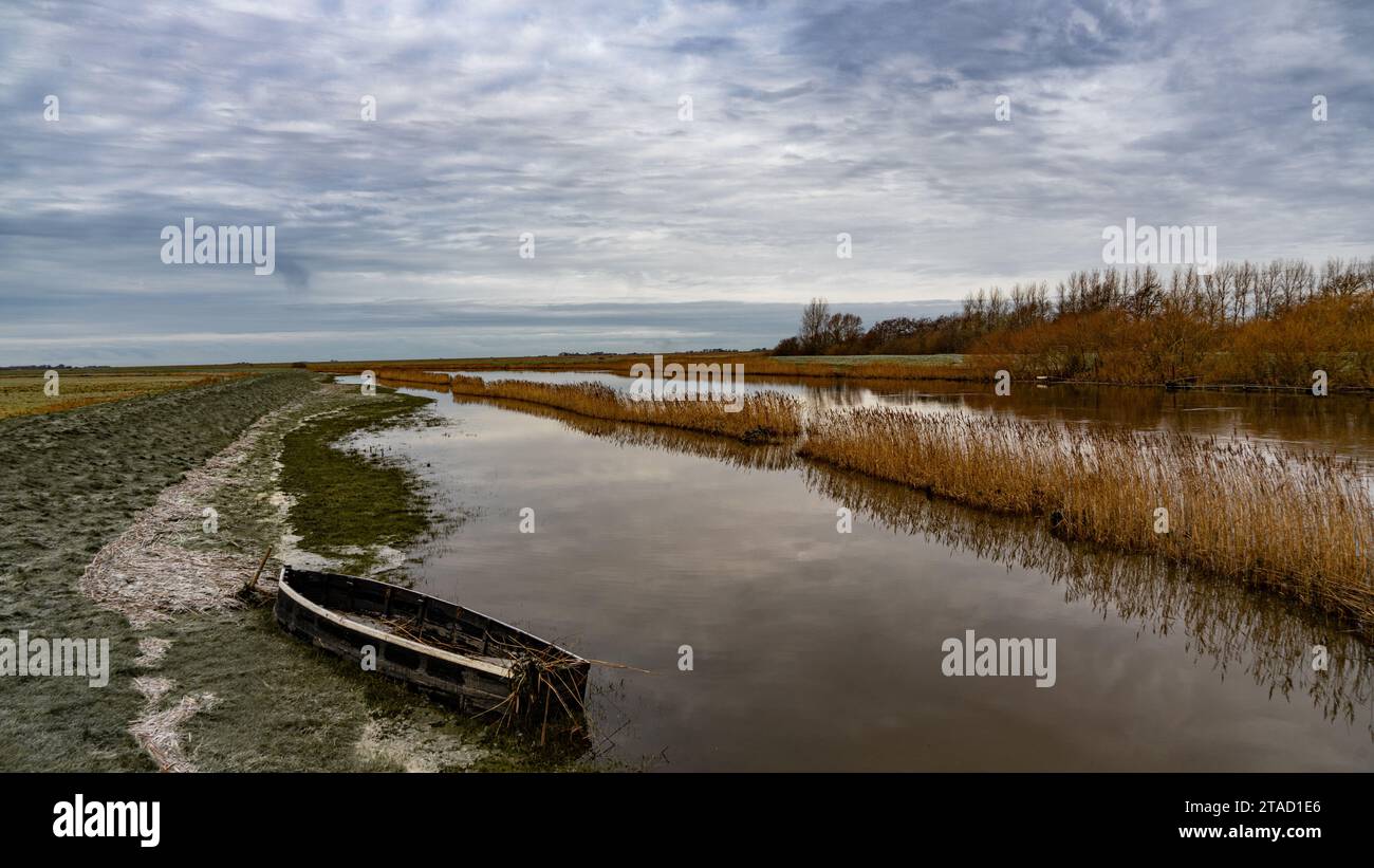 Low clouds hanging over a small river with an old boat on its bank that runs through fields in the Wadden Sea National Park Stock Photo