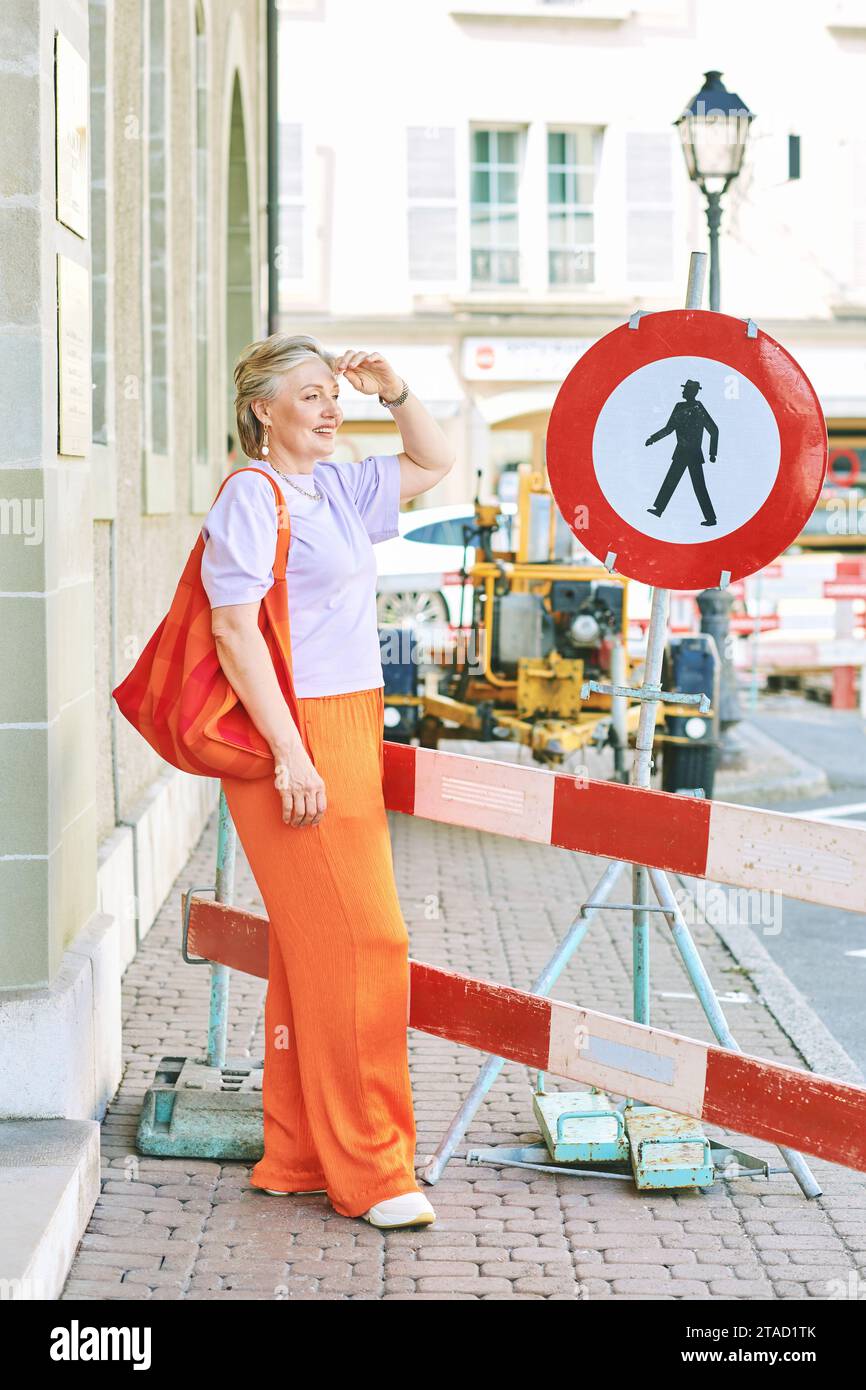 Outdoor fashion portrait of stylish mature woman with grey hair, wearing violet top and orange pants Stock Photo