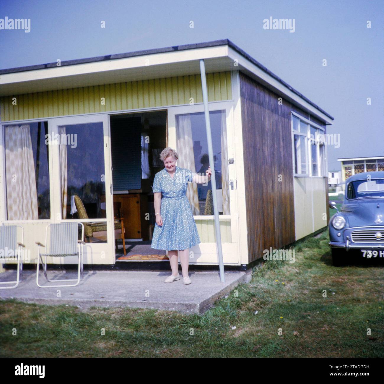 Woman standing in doorway of holiday chalet with Morris Minor car parked, Miami Beach private holiday estate, Sutton on Sea, Lincolnshire, England, UK, 1968 Stock Photo