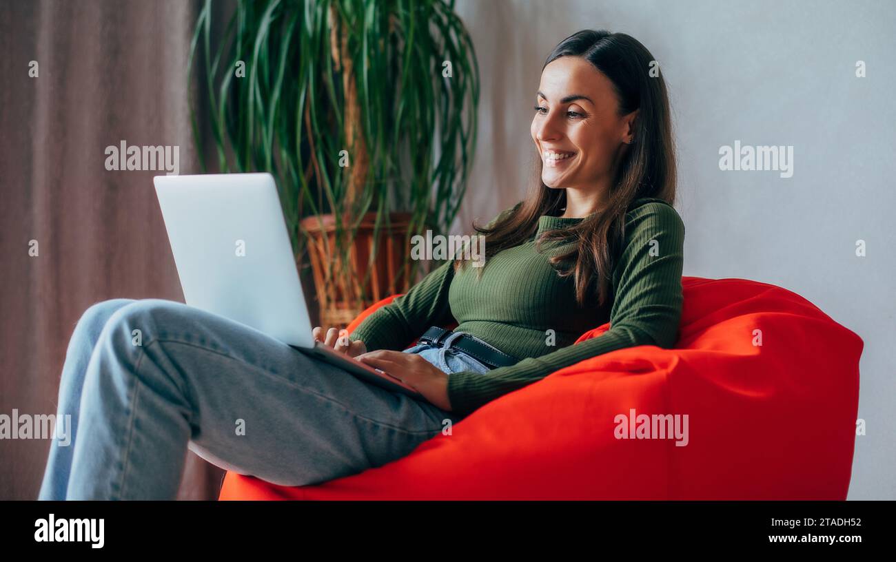 Beautiful young brunette woman using laptop at home while sitting on chair smiling. Freelance, lifestyle concept, rest, relax Stock Photo