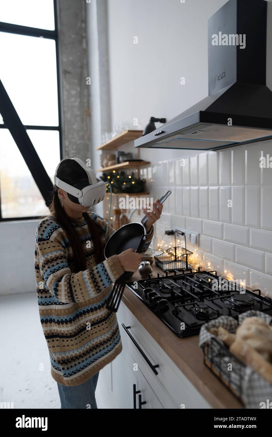 In the festive kitchen, a beautiful and stylish girl is cooking following a recipe with the aid of a virtual reality headset. Stock Photo