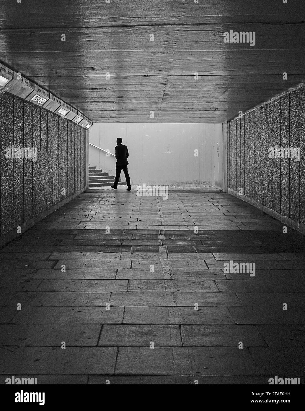 Black and white image of Silhouette of a male person walking in a dark pedestrian underpass towards steps and daylight. Stock Photo