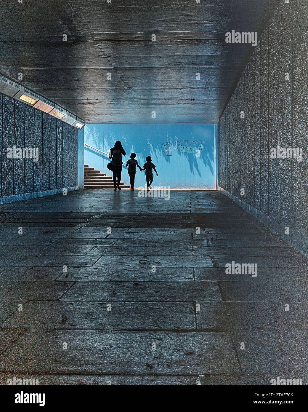 Silhouette of a female & 2 children walking in a dark pedestrian underpass towards steps and daylight. Stock Photo