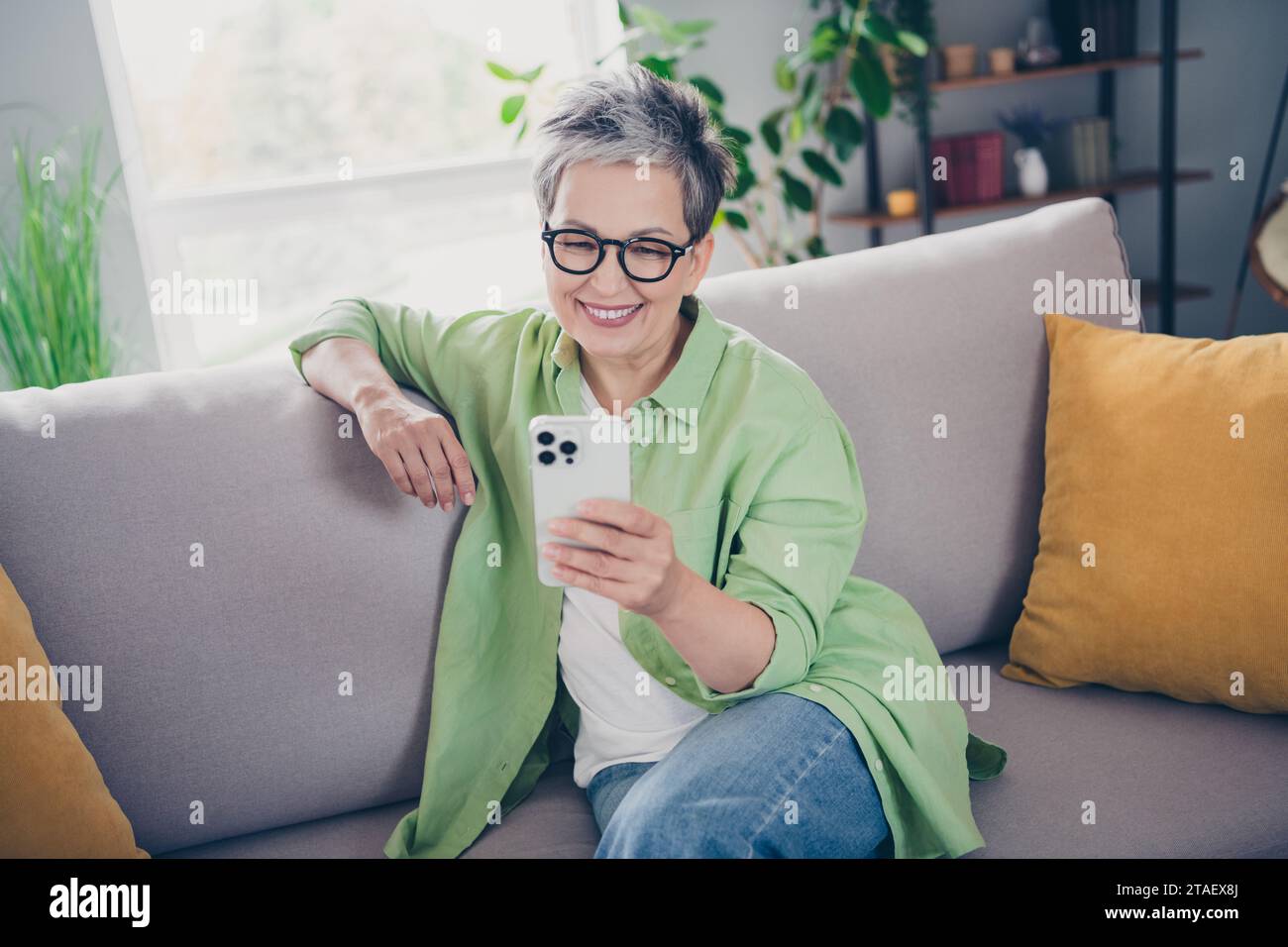 Portrait of beautiful mature age business woman chatting with her grandkids using smartphone sit couch indoors at home modern loft interior Stock Photo