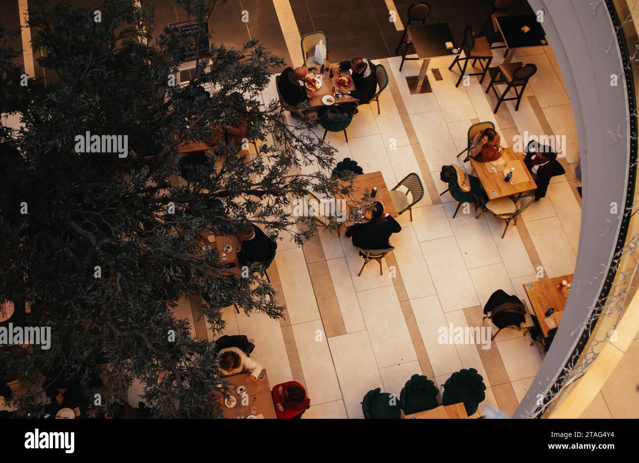 Madrid, Spain. 1 May 2023 Many people sitting eating in food court, cafe, restaurant in shopping mall view from above. Round stairwell. A big tree in Stock Photo