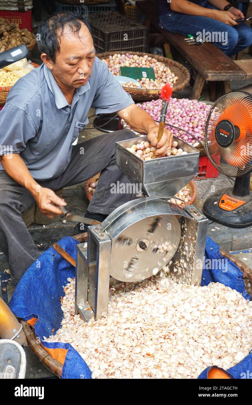 A man uses a hand-cranked slicer to chop garlic cloves at an outdoor market in Hanoi Vietnam. The simple machine facilitates an otherwise tedious task Stock Photo
