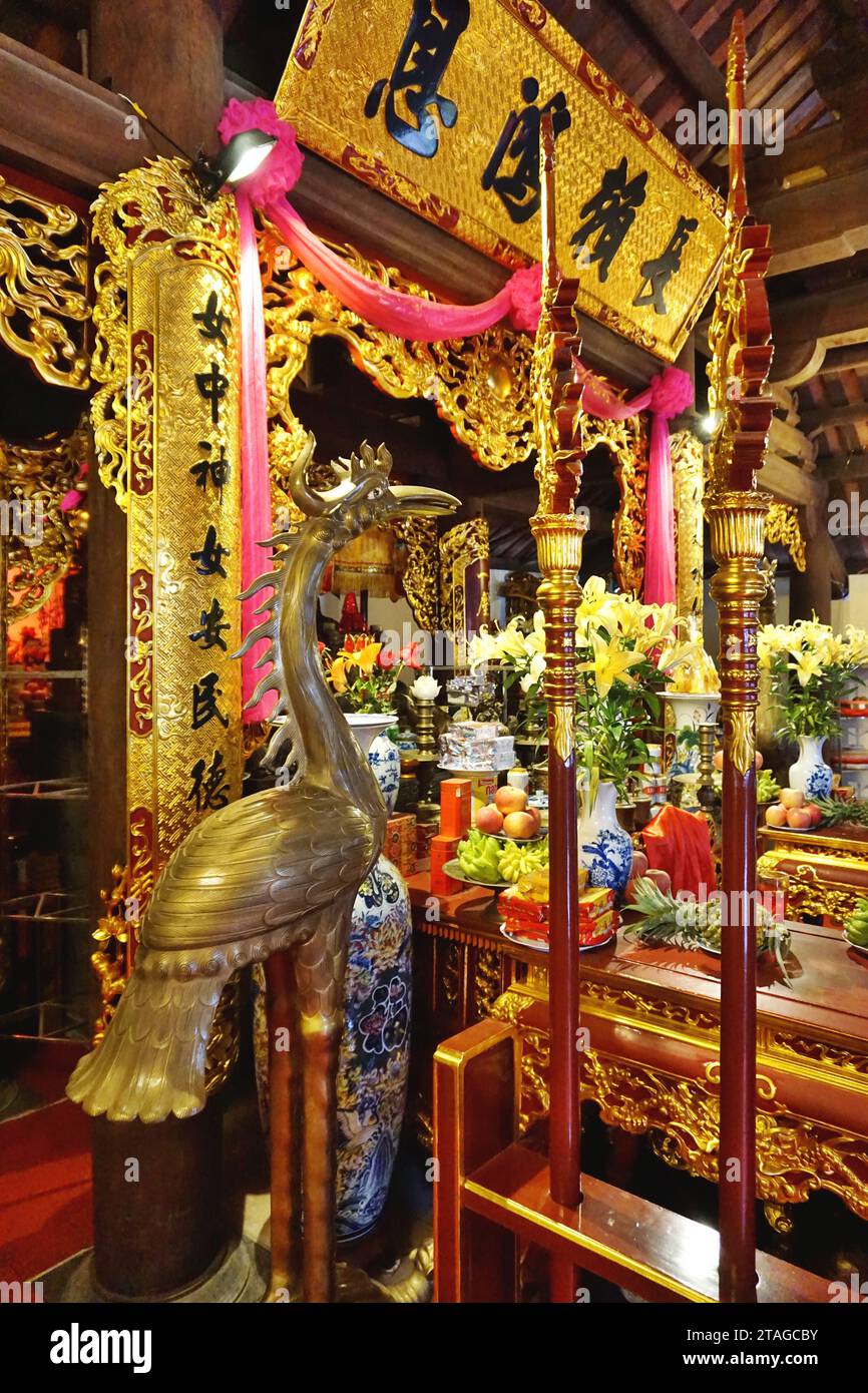 Decorative bronze standing bird looks toward an altar piled high with colorful prayer offerings inside a Vietnamese Buddhist temple in Hanoi, Vietnam Stock Photo