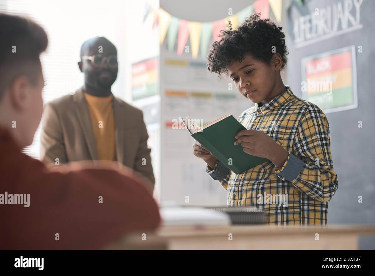 African American schoolboy reading a book to his classmates and teacher ...
