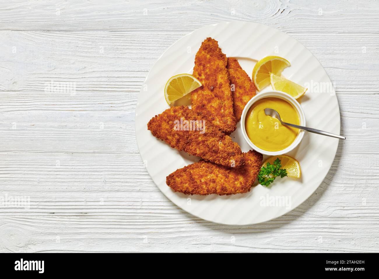 baked in oven breaded fish fillet served with yellow mustard and lemon slices on white plate on white wooden table, flat lay, free space Stock Photo