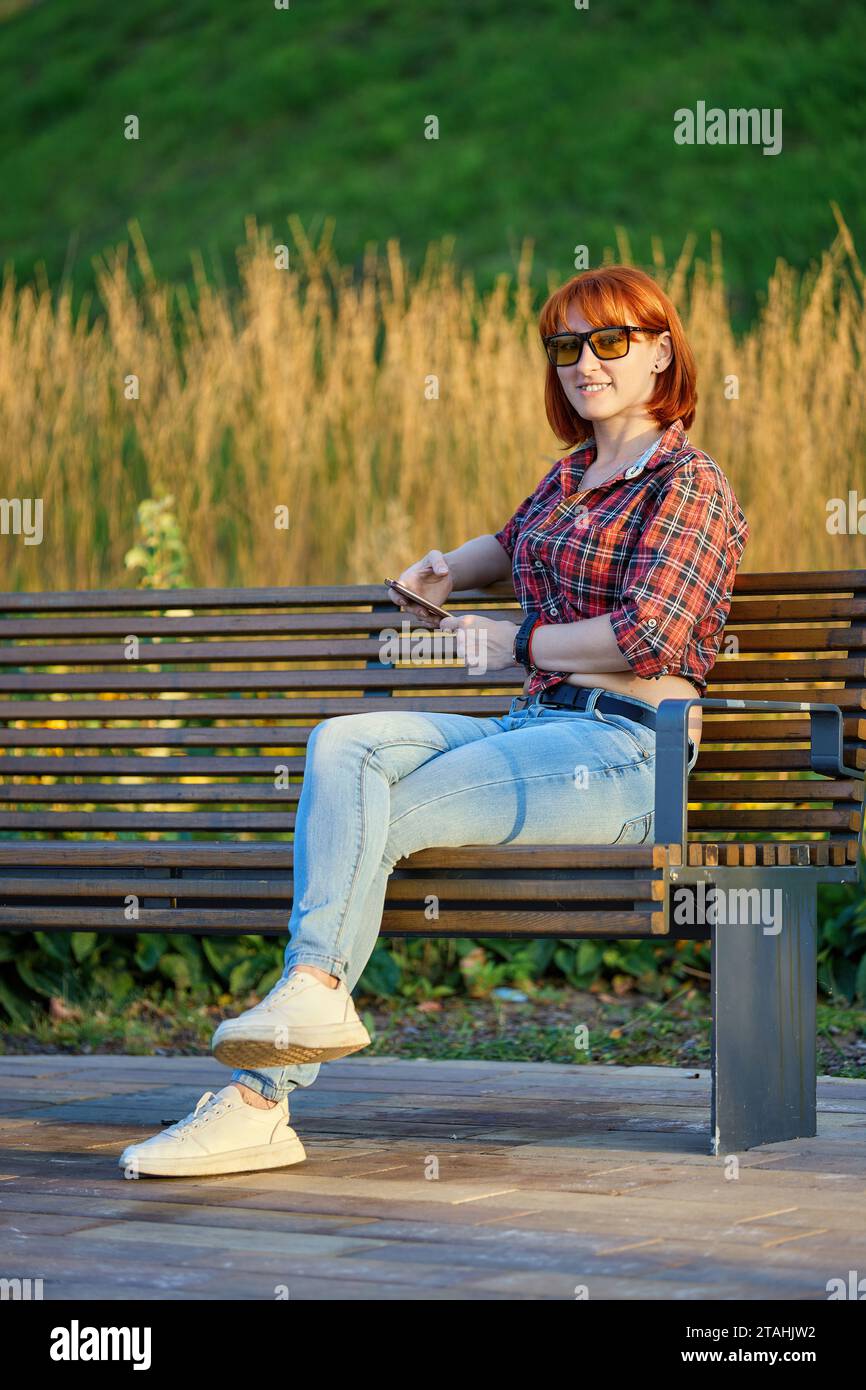 A fashionable redhead woman enjoying an autumn evening in the city. She's wearing a trendy red shirt and denim jeans, making a bold fashion statement Stock Photo