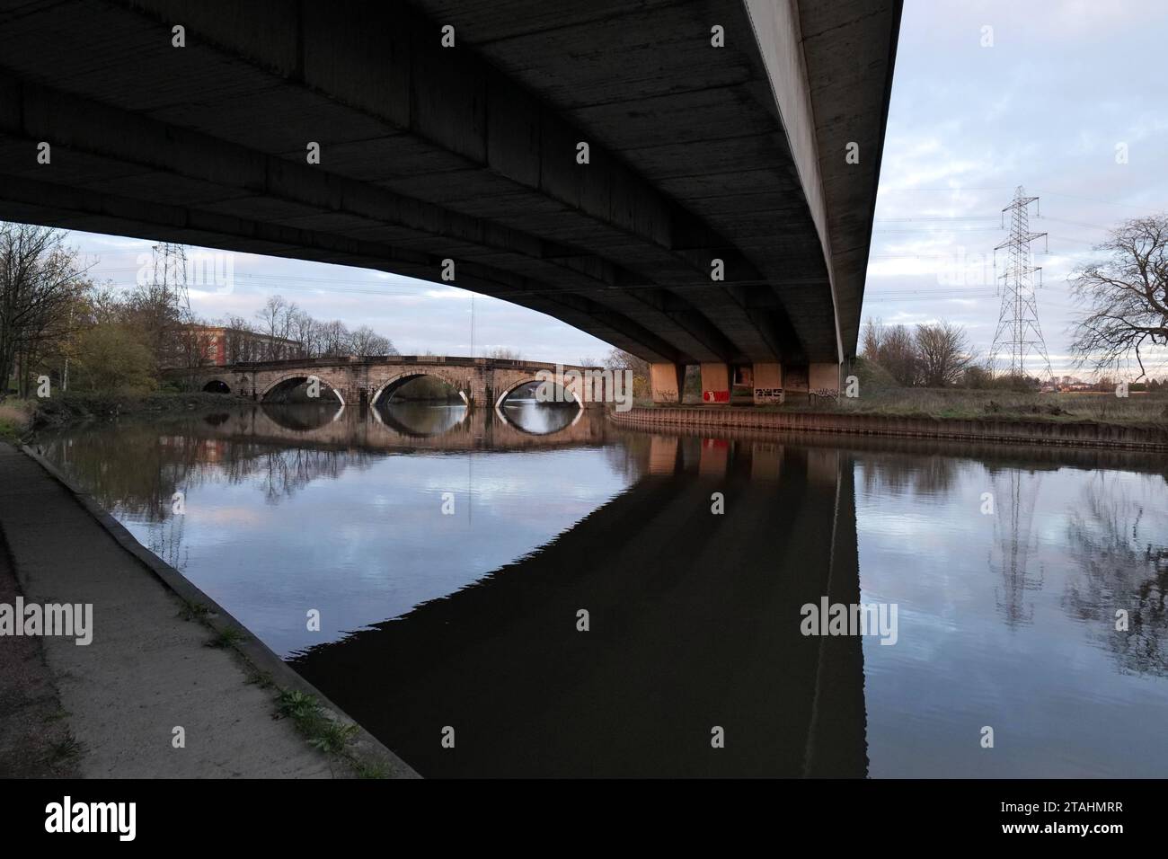 View from underneath a modern dual carriageway road, over the river Aire in UK. An old sandstone bridge can be seen in the background along with elect Stock Photo