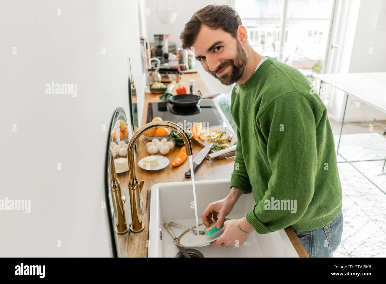 Smiling bearded man in casual clothes looking at camera while washing plate at home Stock Photo