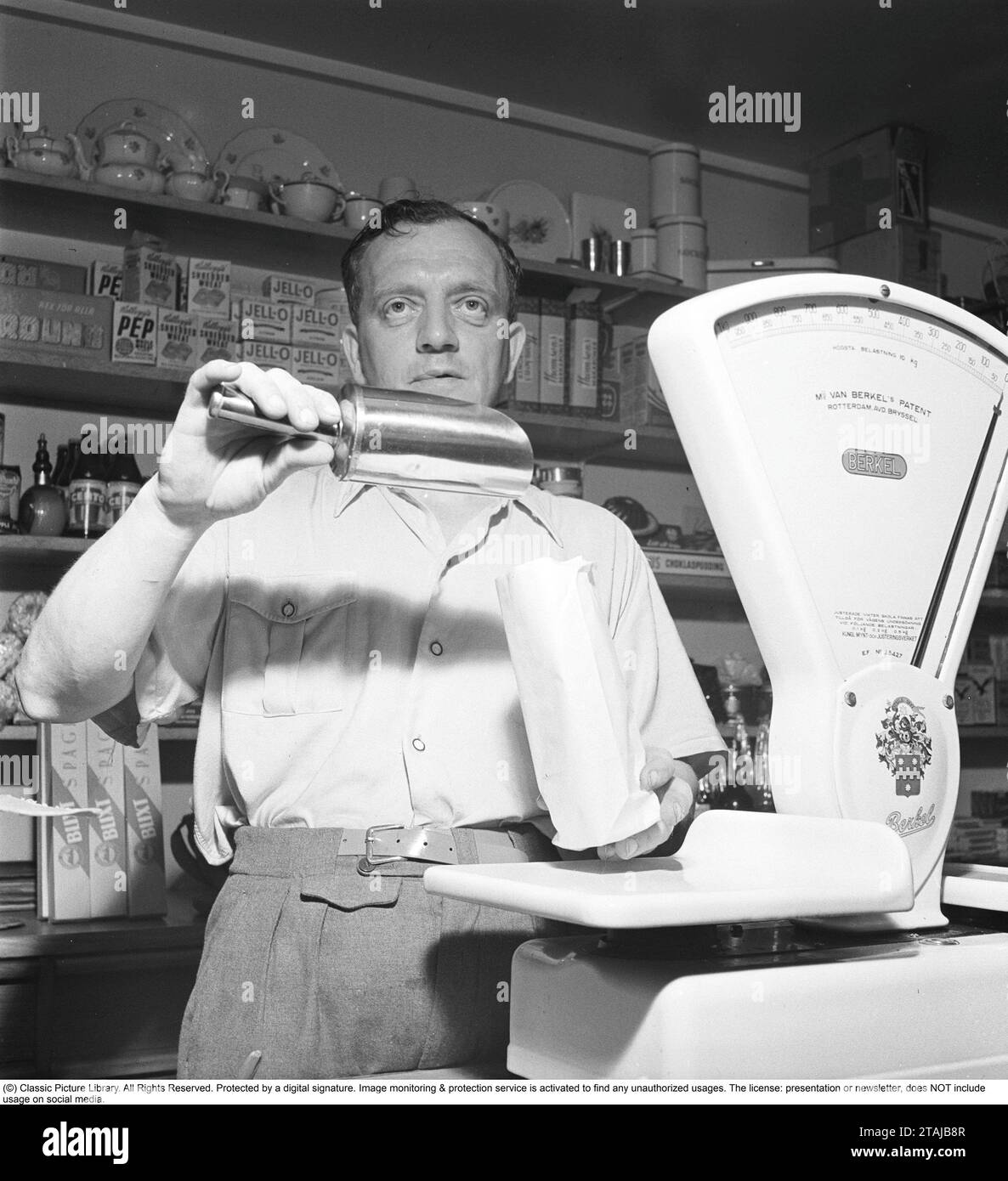 In a grocery store in the 1940s. A shopkeeper is seen pouring something into a paper bag. On the shelves behind him, packages and bottles are neatly lined up. A white shop scale is on the counter and it was an indispensable part of the business as many goods were sold in bulk. 1947. Kristoffersson ref AC127-4 Stock Photo