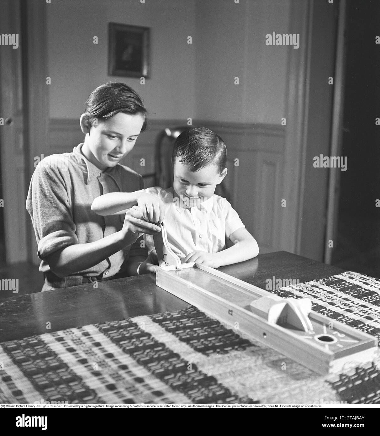 Children play games in the 1940s. In the absence of television and the Internet, the children of the 1940s spent their free time, among other things. with playing board games. Here, two boys playing a game where the aim is to get a coin right along a wooden track. It is dropped and rolls along a track and the goal is to get the coin to stop on a part of the track that gives the highest score. 1949. Kristoffersson ref AU62-5 Stock Photo
