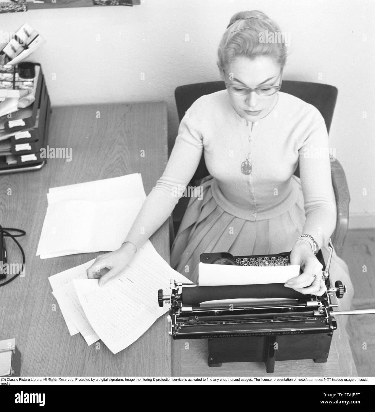 Office girl in the 1950s. A woman is sitting at a office desk typing on her typewriter. 1957. Stock Photo