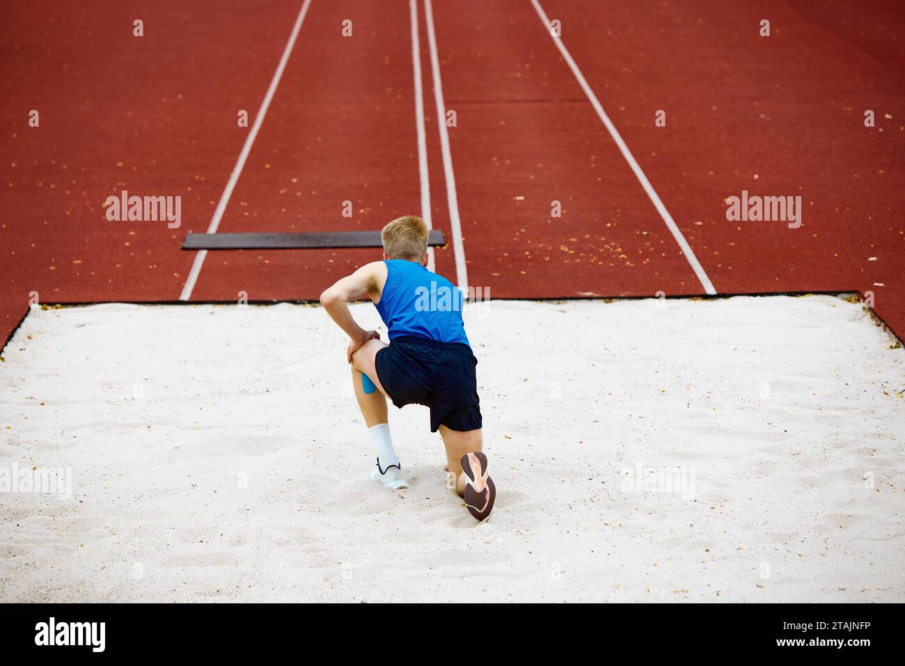 Rear view of young athletic man, professional sportsman wearing in sport uniform stands in sand pit. Intensity captured in mid-stride long jump. Stock Photo