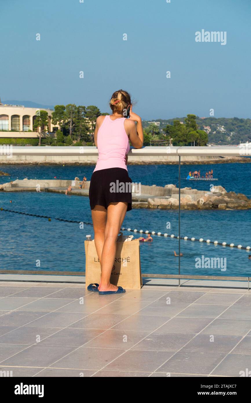 Young woman lady talking on phone / telephone talk / mobile device on Larvotto Promenade, overlooking beautiful Larvotto Beach. Sunny day, Monaco. (135) Stock Photo