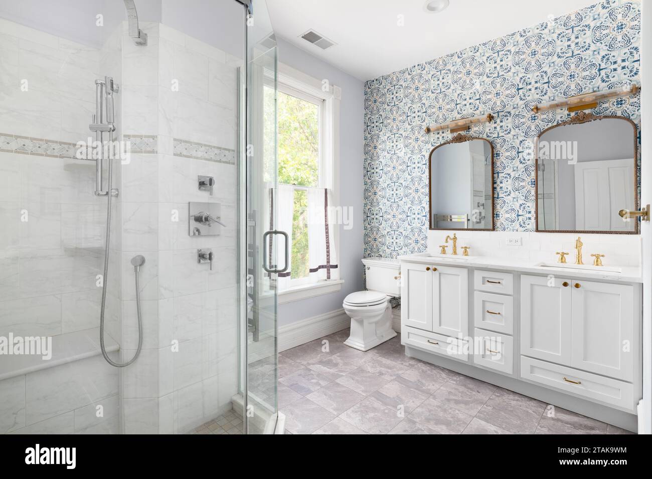 A cozy bathroom with a patterened blue wallpaper, grey cabinet, gold faucet and mirrors, and a tiled walk-in shower. Stock Photo