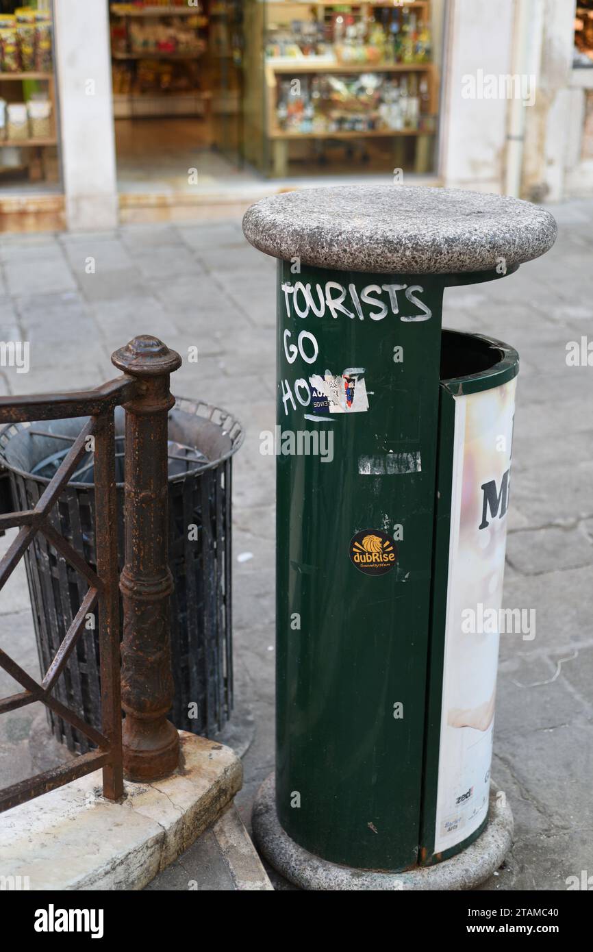 Signs of Overtourism overrunning Venice and good manners – Tourists Go Home, graffiti painted on a green rubbish bin by a popular bridge and shops Stock Photo