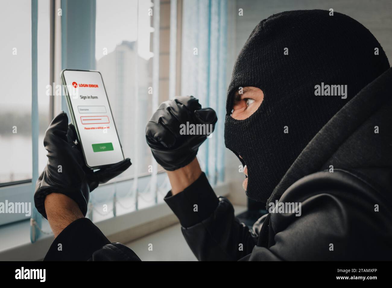 Angry young man in balaclava using cell phone. Theft of personal data. Hacking the city. A bandit in the office picks up a password to log in to a sma Stock Photo