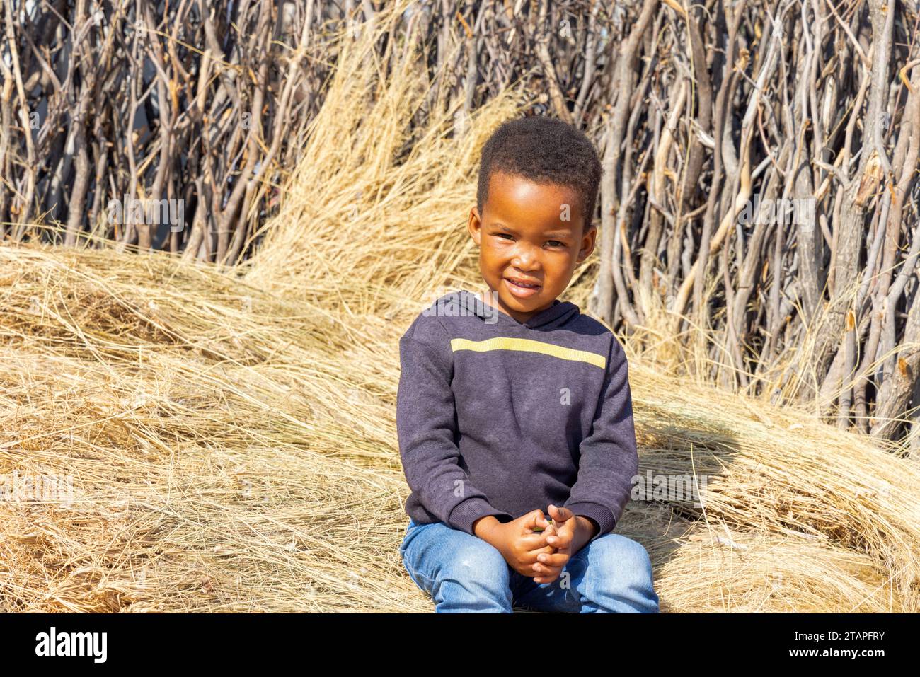 african village , child sitting on thatched grass in the yard by the fence Stock Photo