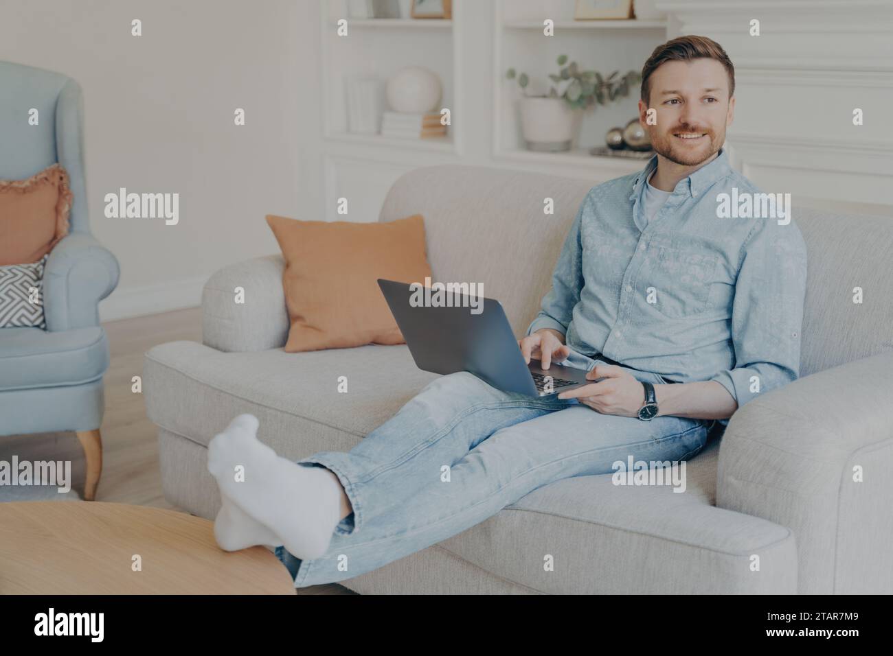 Smiling man working on laptop while sitting comfortably on a sofa in a cozy living room Stock Photo