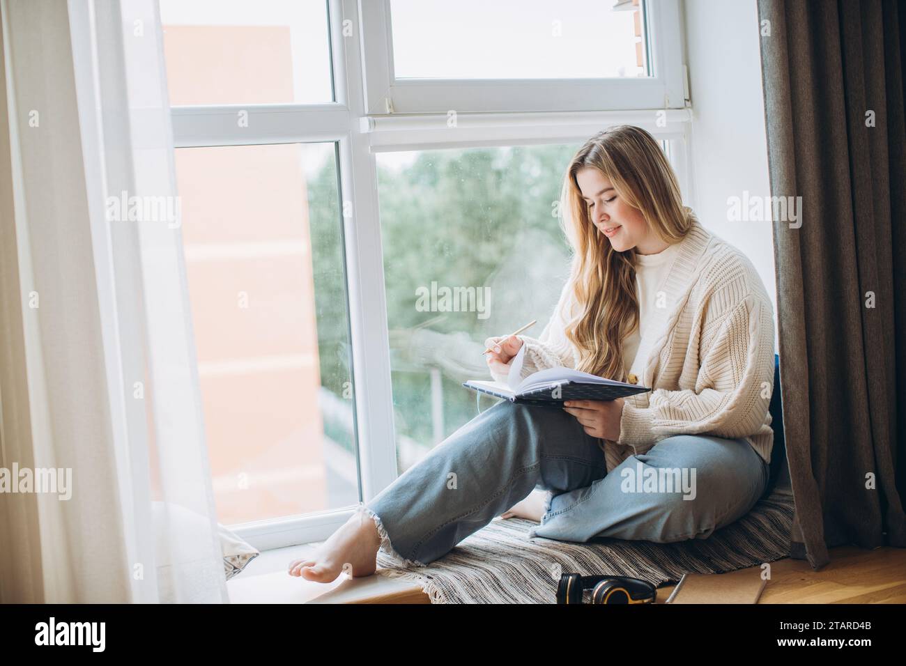 Creative young woman writes memory plan and idea in notebook while sitting near window at home. Creative journal or diary concept. Stock Photo