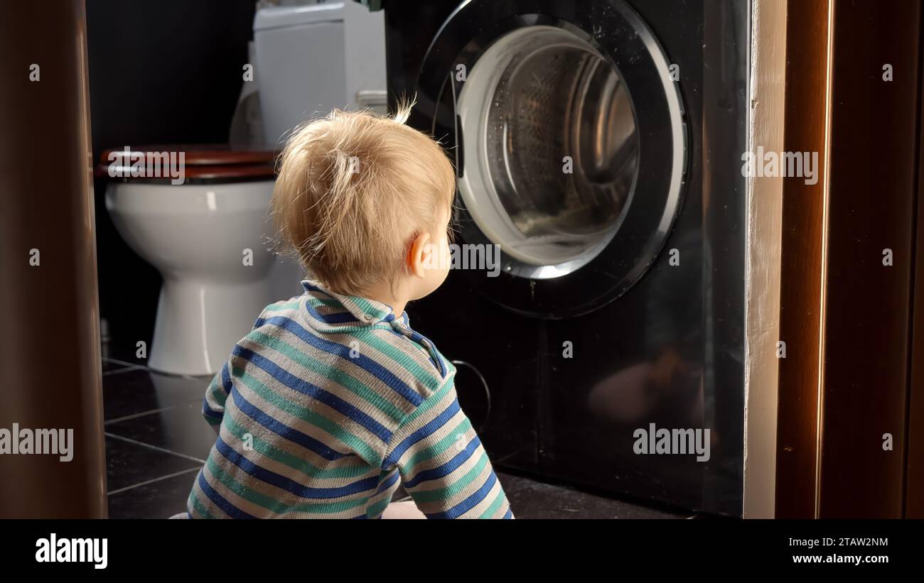 Cute smiling baby boy looking at spinning clothes in washing machine. Doing housework and chores