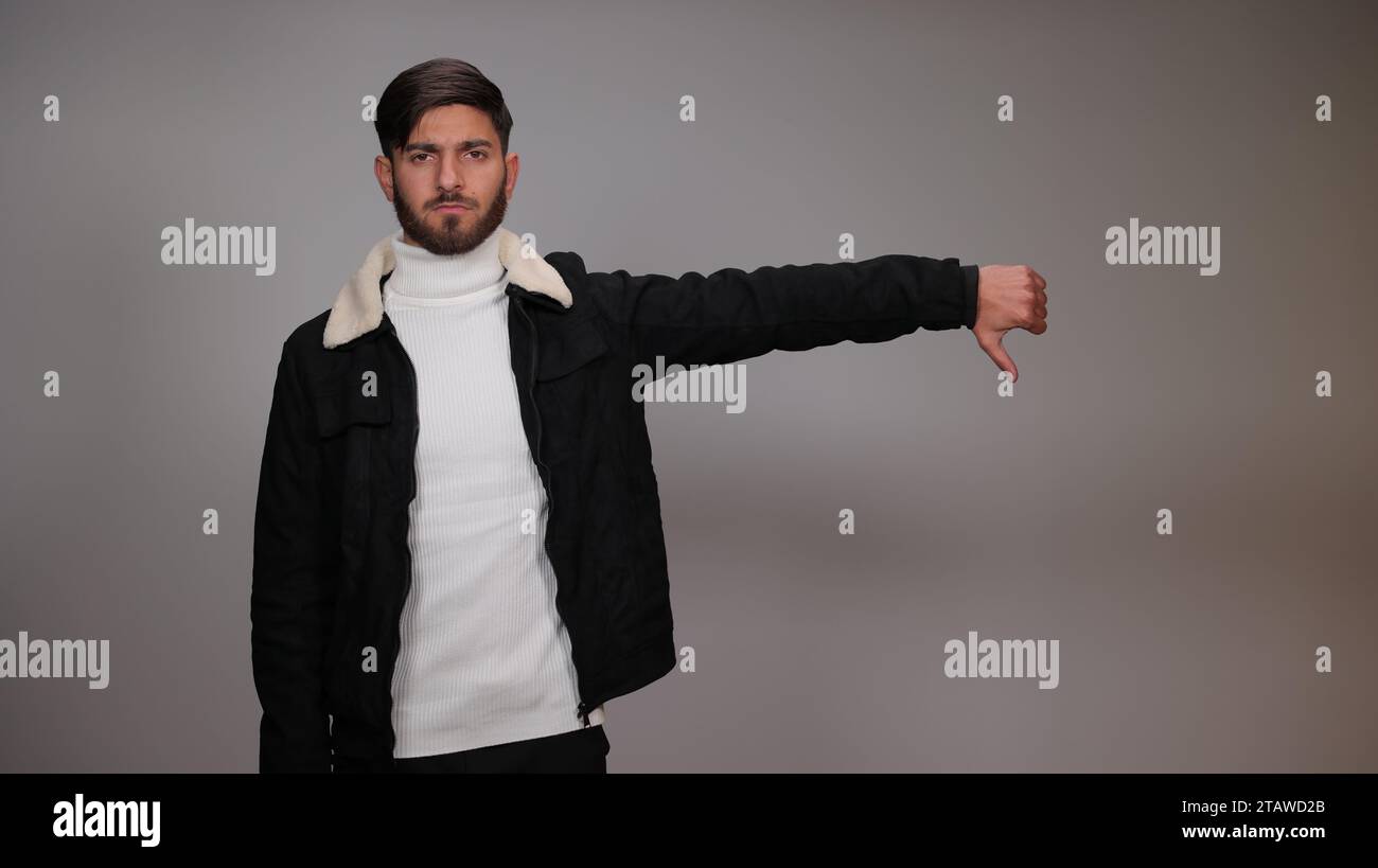 A young man posing with a thumbs-down gesture and expressing dislike against a gray background. Stock Photo