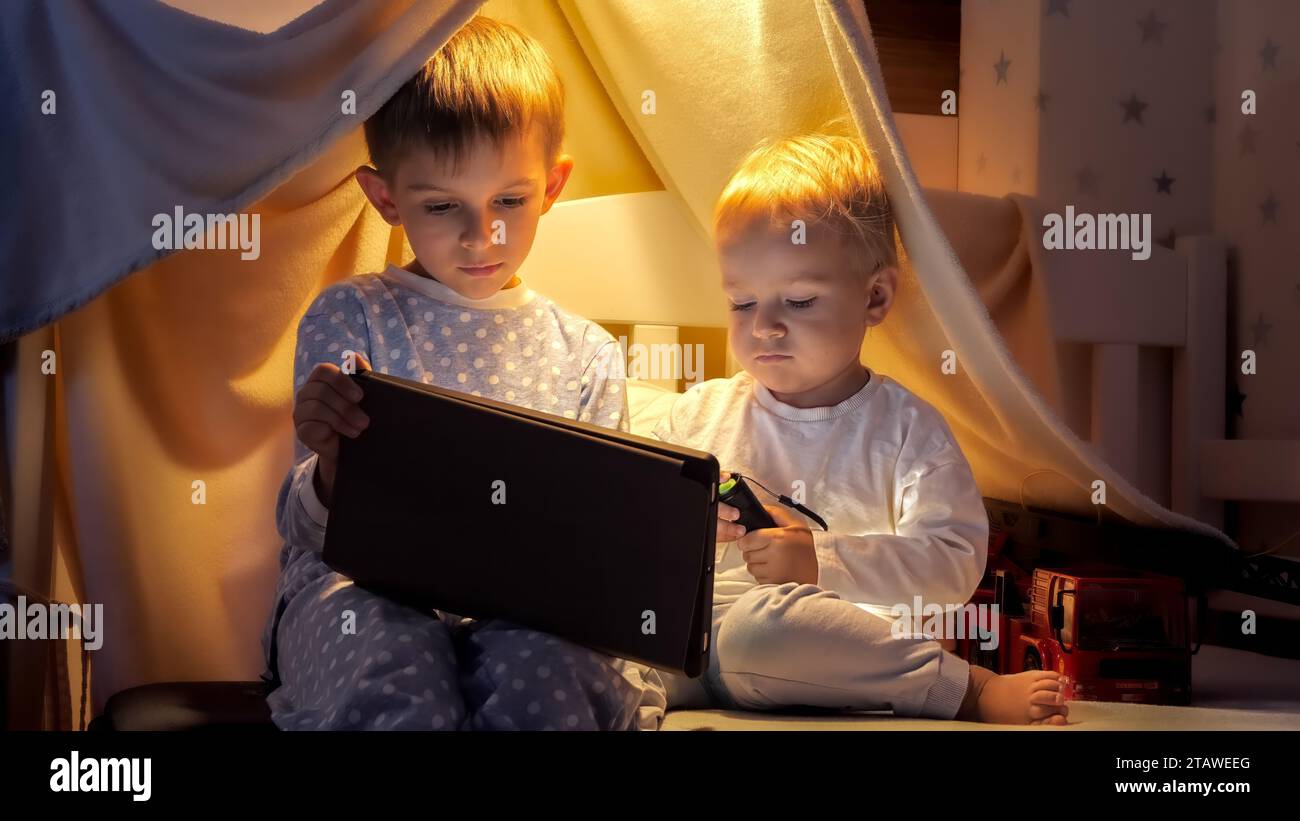Little baby boy with older brother using tablet computer and watching video in bed before gong to sleep at night. Stock Photo