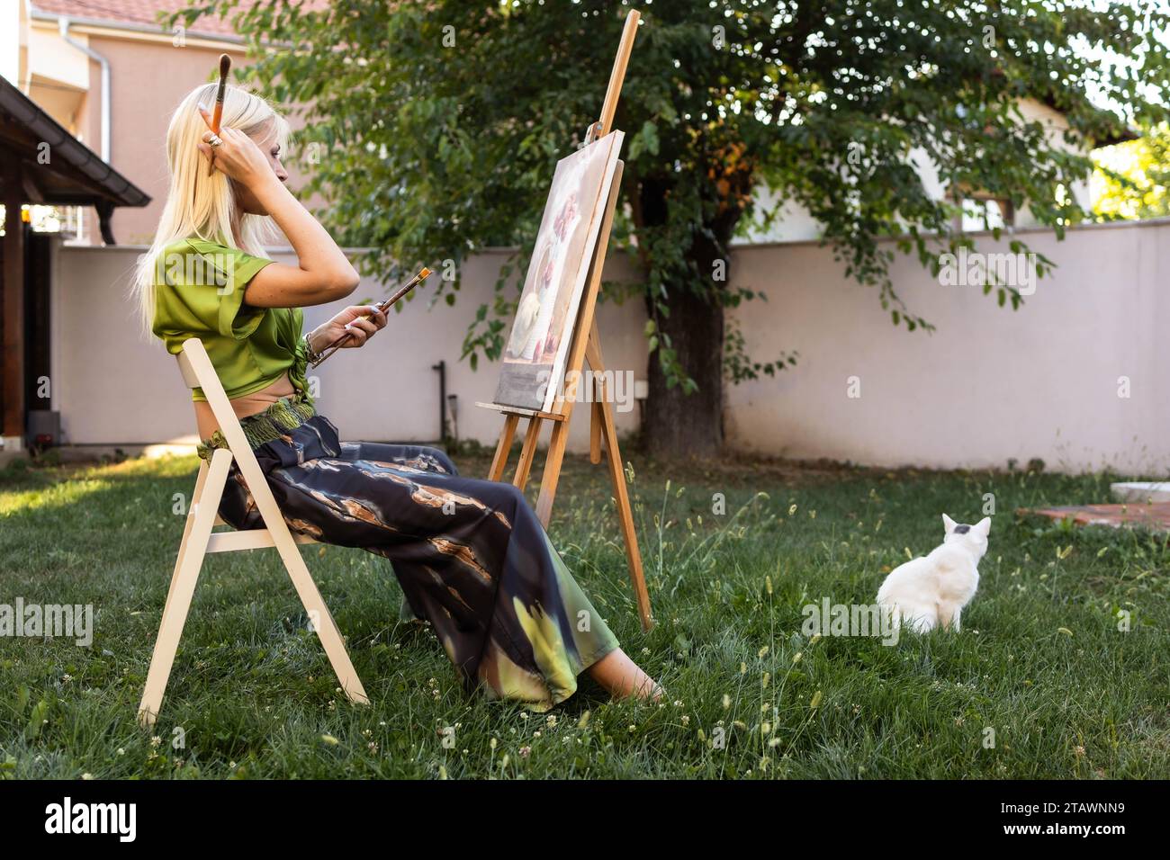 Young, blonde woman in green painting outdoors with cat sitting beside her Stock Photo